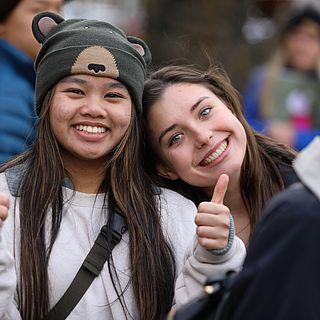 Two female students sit next to each other, smiling and laughing. The student on the right is giving a double thumbs up.